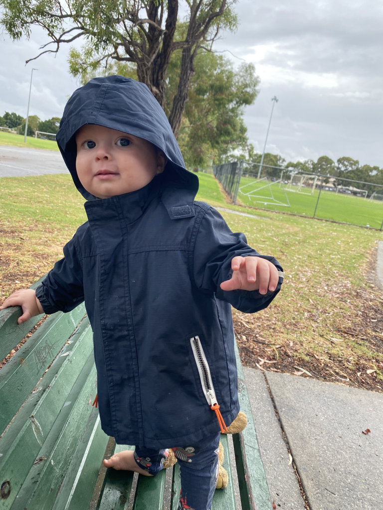 Hugo (17 months old) standing on a park bench outdoors with a rain coat on