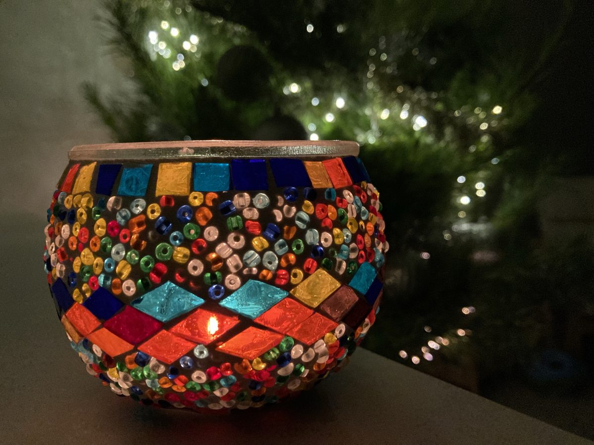A photo of a small candle in a mosaic glass jar, with a christmas tree lit up in the background.